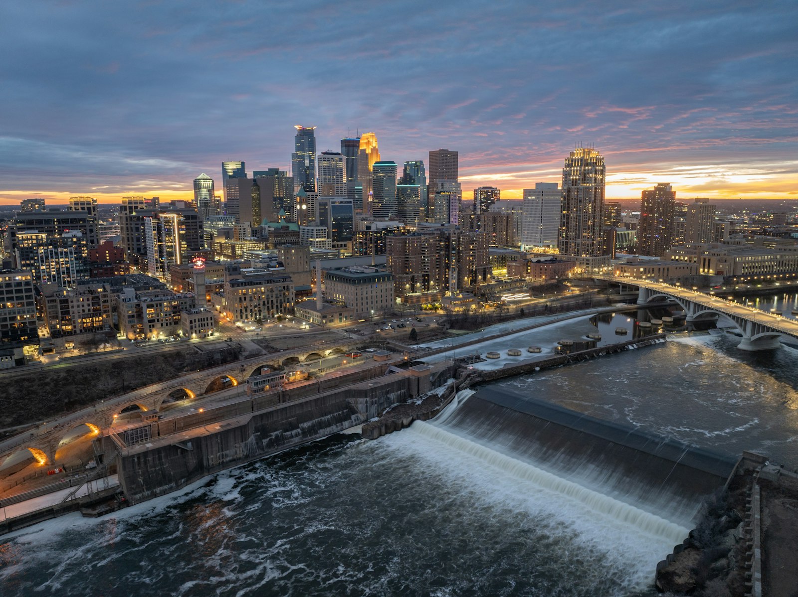 Minneapolis at dusk: A photo of the Minneapolis skyline, taken from the east side of the Mississippi River, with downtown buildings in the background and the Stone Arch Bridge and St. Anthony Falls in the foreground.