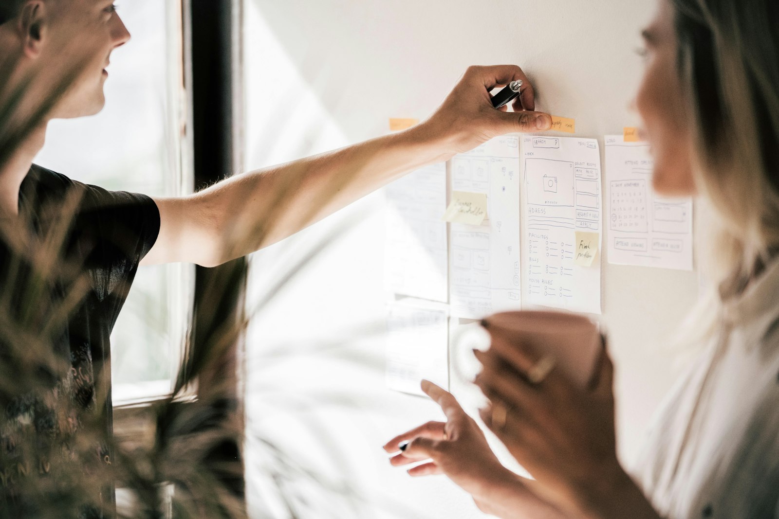 Two smiling people in an office setting converse in front of a pinboard with hand-drawn mockups of webpage layouts. One of the workers affixes a label to one of the mockups, while the other points toward it. Collaboration, design, branding concept.