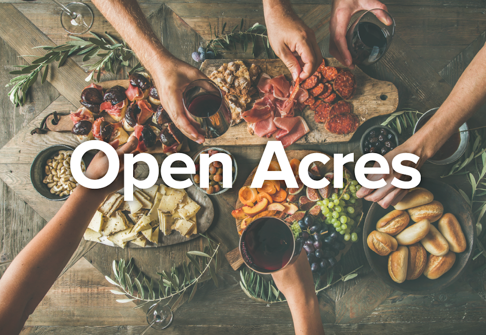 The Open Acres brand name design with white type on photo of a table of food with peoples arms.