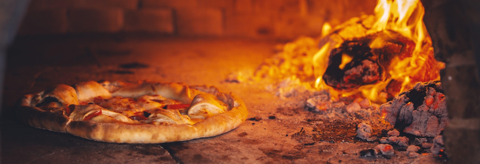 A promotional photograph of pizza going into a wood fire oven.